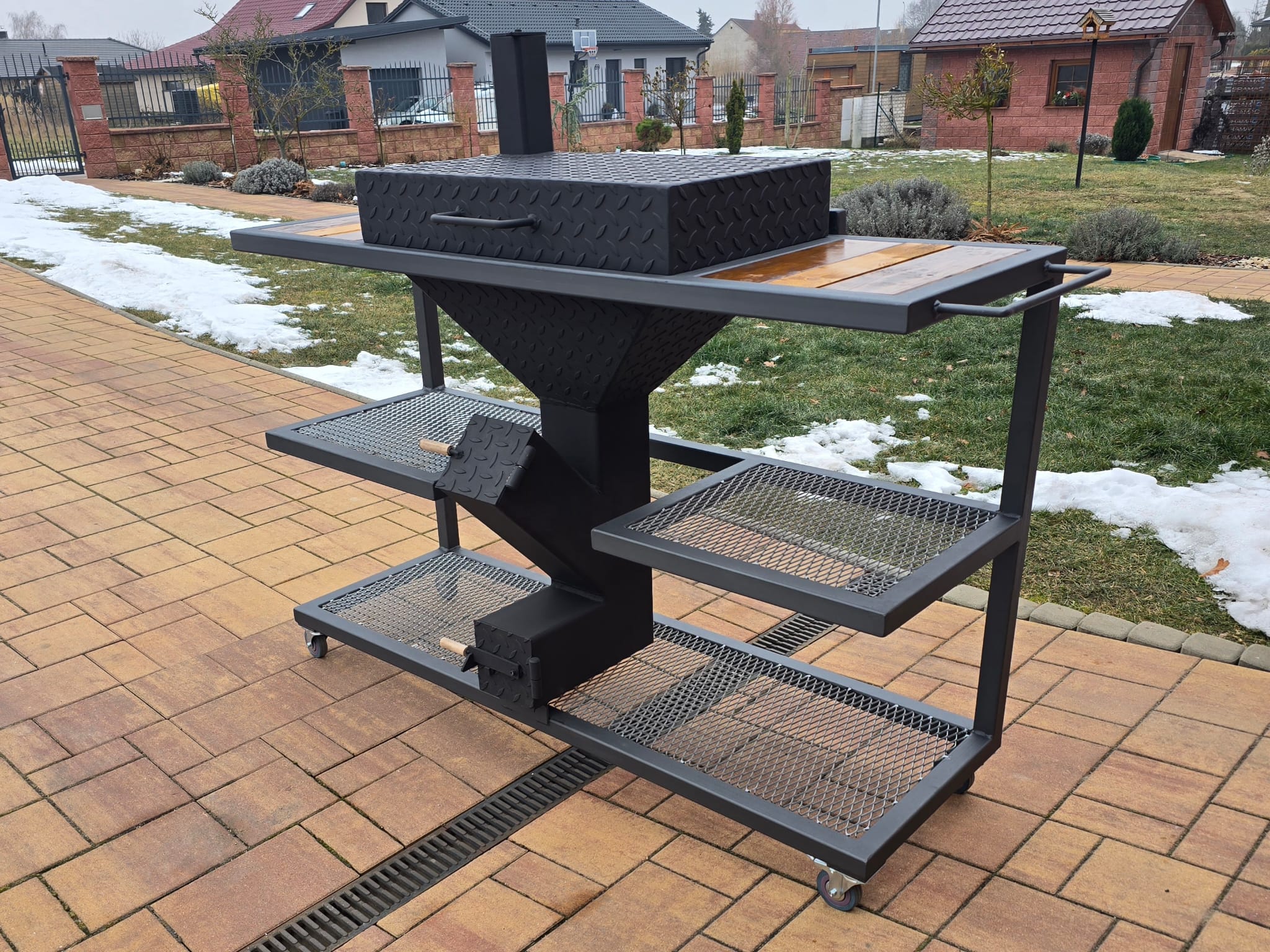 Černý gril s komínem a dřevěnými deskami na terase, víko zavřené